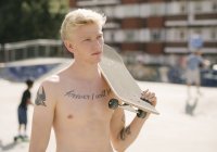 Tattooed young man carrying skateboard on shoulder in skatepark — Stock Photo