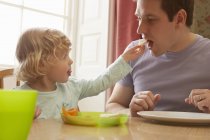 Жінка малюк годує хлібом батька за кухонним столом — стокове фото