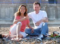 Couple sitting on shingle beach having picnic looking at camera smiling — Stock Photo