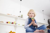 Porträt eines süßen Mädchens, das auf dem Küchentisch sitzt und bunte Karotten in der Hand hält — Stockfoto