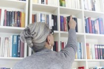Старшая женщина берет книгу с книжной полки, вид сзади — стоковое фото