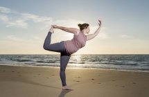 Mulher madura praticando ioga em uma praia ao pôr do sol, pose de árvore — Fotografia de Stock