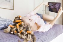 Молода жінка з пінними валиками в волоссі, лежить на ліжку, використовуючи цифровий планшет — стокове фото