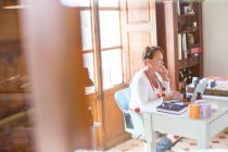 Mulher falando no smartphone enquanto usa laptop no escritório de oficina de sabão artesanal — Fotografia de Stock
