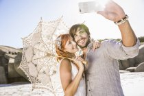 Пара на пляжі тримає мереживні парасольки, використовуючи смартфон, щоб взяти селфі — стокове фото