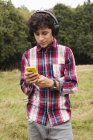 Мальчик-подросток, стоящий в поле, в наушниках, используя смартфон — стоковое фото