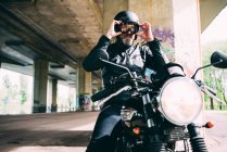 Зрелый мотоциклист, сидящий на мотоцикле, надевает очки под эстакадой — стоковое фото