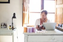 Жінка розмовляє на смартфоні, використовуючи ноутбук в офісі мильної майстерні ручної роботи — стокове фото
