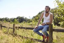Середній дорослий чоловік сидить на сільському паркані, дивлячись на — стокове фото