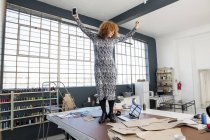 Diseñadora de moda femenina madura bailando con música para teléfonos inteligentes en la mesa del taller - foto de stock