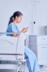 Жінка-лікар, що сидить на лікарняному ліжку, читає текстові повідомлення смартфона — стокове фото