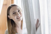 Porträt einer Frau, die hinter den Vorhang blickt und in die Kamera lächelt — Stockfoto