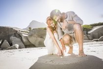 Couple crouching on beach drawing heart shape in sand — Stock Photo