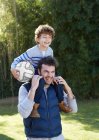 Padre llevando sonriente hijo sosteniendo fútbol sobre hombros - foto de stock