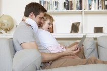 Женщина-ребенок сидит на диване с отцом, глядя на цифровой планшет — стоковое фото
