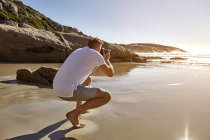 Зрелый человек, приседающий на пляже, фотографирующий вид, Кейптаун, Южная Африка — стоковое фото