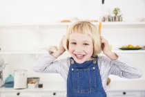 Portrait of cute girl in kitchen holding carrots to her ears — Stock Photo