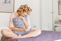Молодая женщина с пенокатками в волосах, сидящая на кровати, обнимающая подушку, используя смартфон — стоковое фото