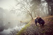 Mulher adulta média agachado com filho criança na margem do rio nebuloso — Fotografia de Stock