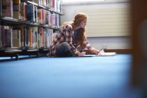 Joven estudiante universitaria escribiendo notas en el piso de la biblioteca - foto de stock