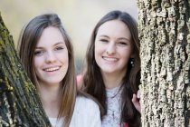 Portrait d'amies derrière des troncs d'arbre regardant la caméra sourire — Photo de stock