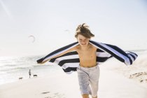 Niño en brazos de playa abierto sosteniendo la toalla mirando a la cámara sonriendo - foto de stock