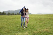 Dos mujeres jóvenes corriendo colina abajo sosteniendo anorak en la lluvia, Sattelbergalm, Tirol, Austria - foto de stock
