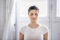Mujer joven meditando con los ojos cerrados en apartamento - foto de stock