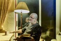 Hombre mayor con libro soñando despierto en la sala de estar al anochecer - foto de stock