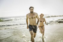 Взрослая пара в бикини и купальных шортах, бегущая в море, Кейптаун, ЮАР — стоковое фото