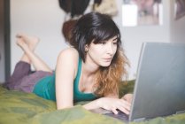 Молода жінка лежить спереду на ліжку, використовуючи ноутбук — стокове фото
