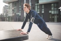 Young male runner doing push ups on bench in city square — Stock Photo