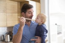 Man feeding toddler son with wooden spoon in kitchen — Stock Photo