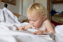 Female toddler lying in bed reading a book — Stock Photo