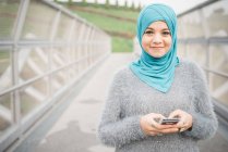 Портрет молодой женщины в бирюзовом хиджабе с помощью смартфона на пешеходном мосту — стоковое фото