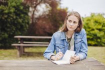 Молода жінка сидить на лавці для пікніка в парку, пишучи в блокноті — стокове фото