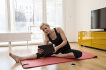 Donna matura seduta su tappetino yoga con gli auricolari che tengono tablet digitale — Foto stock
