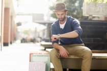Mittlerer erwachsener Mann sitzt auf Sitz, schaut auf Uhr, Einkaufstaschen neben sich — Stockfoto