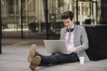 Young businessman sitting on sidewalk and using laptop — Stock Photo