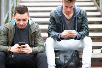 Deux jeunes hommes, assis sur des marches, utilisant des smartphones, à l'extérieur — Photo de stock