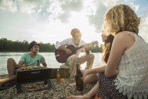 Молодой человек сидит у озера с друзьями, играющими на гитаре — стоковое фото