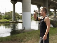 Молодой человек у реки, пьет из бутылки с водой — стоковое фото