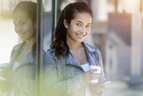 Portrait of young woman with takeaway coffee and smartphone leaning against park building — Stock Photo