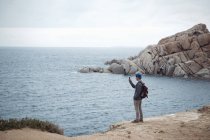 Giovane in piedi sulla scogliera con lo smartphone per fotografare l'oceano, Costa Smeralda, Sardegna, Italia — Foto stock