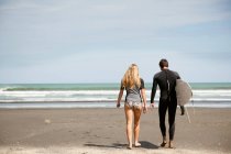 Pareja joven caminando hacia el mar, joven llevando tabla de surf - foto de stock
