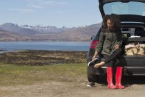 Mujer adulta sentada en una bota de auto poniéndose zapatos, Loch Eishort, Isla de Skye, Hébridas, Escocia - foto de stock