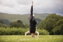 Зріла жінка практикує йогу стоячи на голові з піднятою ногою в полі — стокове фото