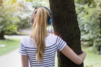 Rear view of young woman with ponytail, wearing headphones, arm around tree looking away — Stock Photo