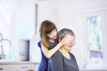 Girl covering grandmothers eyes at kitchen table — Stock Photo
