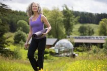 Mujer madura llevando esterilla de yoga caminando en el campo eco lodge - foto de stock